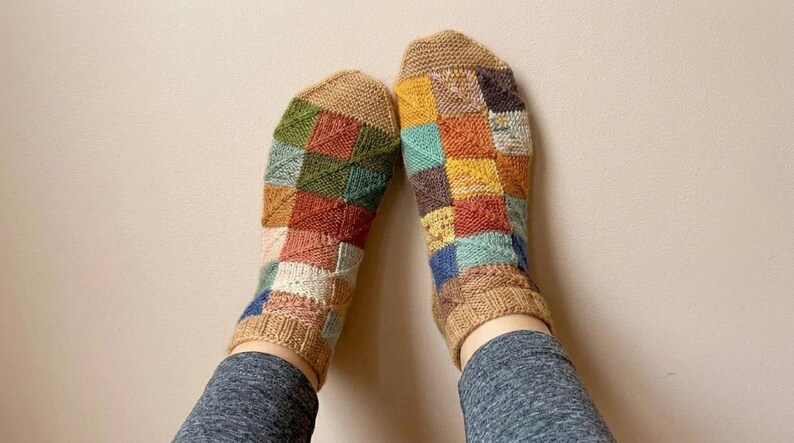 Pode incluir: Um par de meias coloridas de malha com um design de patchwork. As meias apresentam quadrados de v&aacute;rias cores, incluindo castanho, verde, laranja, azul e amarelo. Os punhos e os dedos s&atilde;o de cor bege lisa.