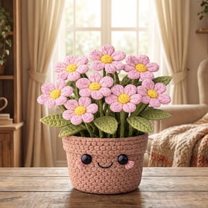 May include: A crocheted flower pot with a smiling face, holding a bouquet of pink and yellow crocheted flowers. The pot is a light brown colour and sits on a wooden table. The background includes a window, curtains, and a wooden shelf.