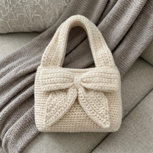 May include: A cream-colored crochet handbag featuring a prominent, large bow detail on the front. The bag is constructed with a dense, textured stitch pattern and includes a single, integrated handle at the top, maintaining a uniform off-white color.