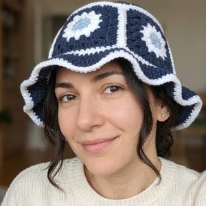 May include: A navy blue and white crocheted bucket hat with a floral design. The hat features a scalloped brim and is made of a soft, textured yarn. The hat is a stylish accessory.