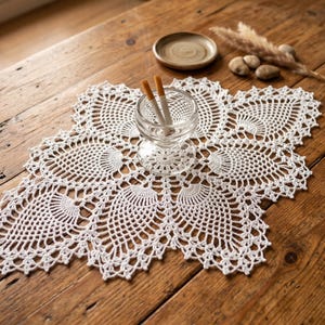 May include: A white, handmade crochet doily with an intricate floral design sits on a rustic wooden table. A small glass ashtray with two cigarettes is centered on the doily. A small ceramic dish and decorative elements are also on the table.