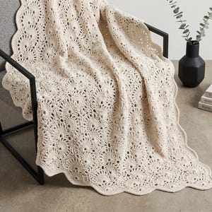 May include: Cream-colored, crocheted blanket with a scalloped edge draped over a gray armchair with a black metal frame. The blanket features a detailed circular pattern. A black vase with greenery and books are in the background.