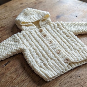 Puede incluir: Cárdigan de bebé color crema con capucha y diseño de punto de cable. El suéter de punto tiene una parte delantera abotonada y puños acanalados. La prenda está hecha de un hilo suave y texturizado, adecuado para bebés.
