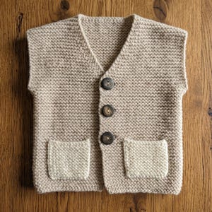 May include: A beige knitted vest with a V-neck, three dark brown buttons, and two front pockets. The vest is made of a chunky knit fabric and is displayed on a wooden surface.