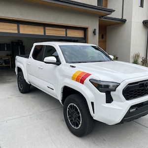 Pode incluir: Caminhonete Toyota Tacoma branca com rodas e acabamentos pretos. Apresenta um decalque de listras coloridas no painel lateral dianteiro. A caminhonete est&aacute; estacionada em uma entrada em frente a uma casa moderna com garagem.