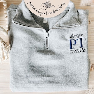 Puede incluir: Sudadera gris con cremallera con el texto "Angie PT PHYSICAL THERAPIST" bordado en la parte delantera en hilo azul.