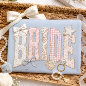 May include: A light blue fabric with the word "BRIDE" appliqued in various patterns, including lace, floral, and gingham. Embellishments include bows, a star, a heart, and a ring. The design is set on a woven basket.