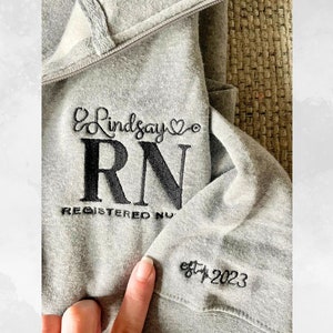May include: Grey hooded sweatshirt with black embroidered text that reads "Lindsay RN Registered Nurse est. 2023".