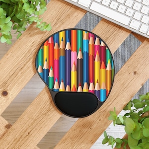 May include: Ergonomic mouse pad with a colorful pencil design. The pad features a black wrist rest for comfort and a vibrant array of colored pencils arranged vertically. The design includes red, yellow, blue, and green pencils.