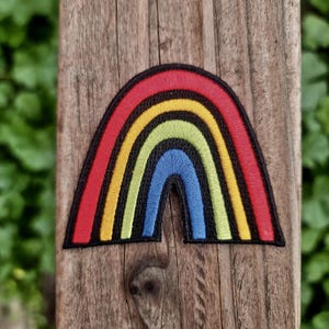 May include: Embroidered rainbow patch with red, yellow, green, and blue arches, outlined in black. The patch is affixed to a weathered wooden surface, with a blurred green background.