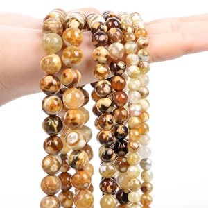 Puede incluir: Una hebra de cuentas de ágata marrón y blanca con un patrón natural de mármol. Las cuentas son redondas y tienen un acabado liso y pulido.