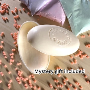 May include: An oval-shaped off-white bar of soap with the embossed text Orchard & Vine, accompanied by scattered small peach-colored wax beads and three pastel-colored pouches. The text Mystery gift included is visible at the bottom.