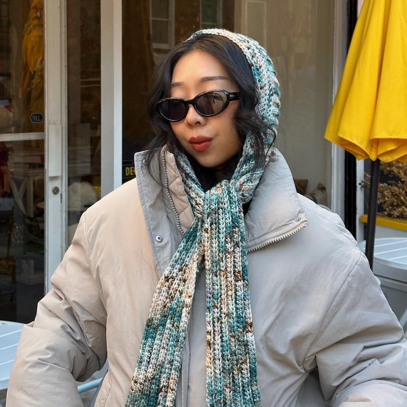 Puede incluir: Un conjunto de gorro y bufanda de punto en tonos turquesa, marr&oacute;n y crema. El gorro es de estilo beanie ajustado, y la bufanda es larga y rectangular. El conjunto se lleva con una chaqueta acolchada de color claro y gafas de sol.