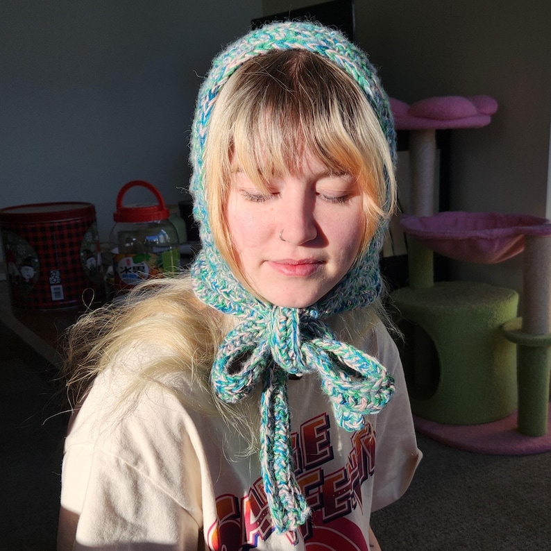 Puede incluir: Un gorro hecho a mano, tejido a ganchillo en tonos de azul, verde y blanco. El gorro presenta un lazo en el cuello y est&aacute; hecho de un hilo suave y texturizado. La persona de la imagen lleva una camiseta blanca con un dise&ntilde;o gr&aacute;fico.