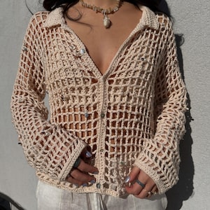 Puede incluir: Un top de crochet beige con cierre de botones en la parte delantera y mangas largas. El top tiene un diseño suelto de tejido abierto y está decorado con pequeñas cuentas plateadas.