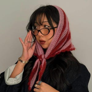 Puede incluir: Un pañuelo de ganchillo a rayas rosas y rojas atado bajo la barbilla. El pañuelo está hecho de un hilo suave y texturizado. La bufanda es un accesorio de moda.