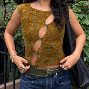 May include: A sleeveless knit top in a mottled mustard yellow and forest green colourway, featuring a diagonal front opening secured by four small teal buttons, creating a series of cutouts along the centre of the garment.