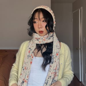 Puede incluir: Un conjunto de bufanda, gorro y bufanda a juego de punto color crema. El conjunto presenta un patr&oacute;n moteado con acentos rojos, azules y grises. La bufanda est&aacute; drapeada alrededor del cuello y el gorro se lleva en la cabeza.