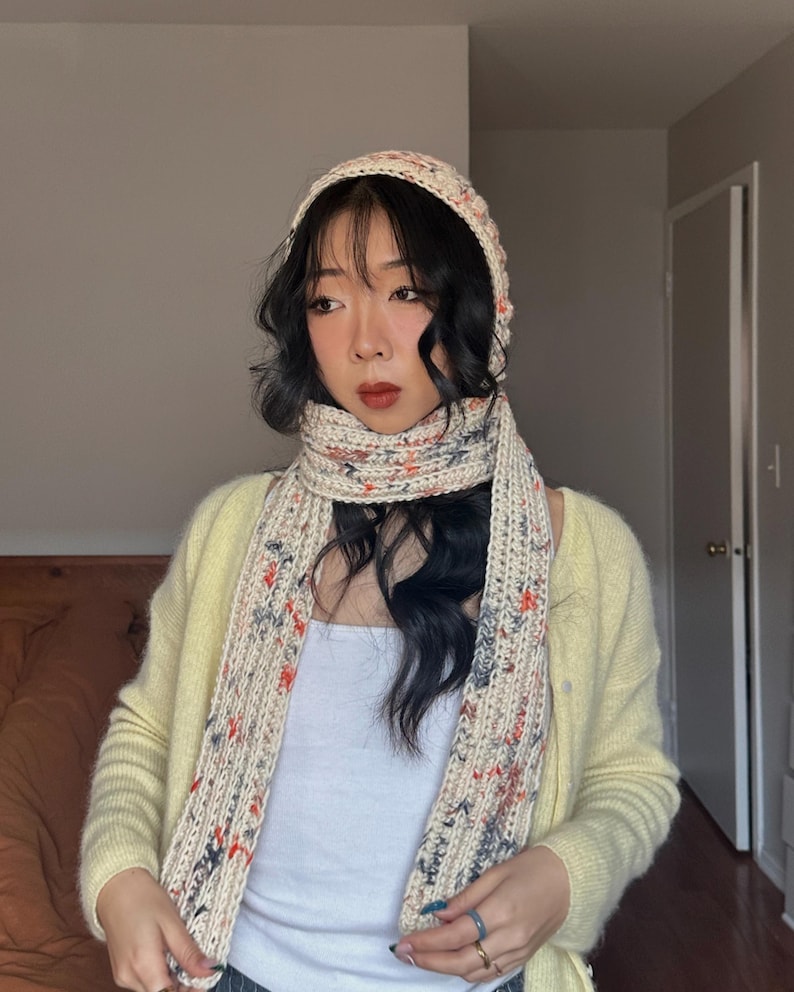 Puede incluir: Un conjunto de bufanda, gorro y bufanda a juego de punto color crema. El conjunto presenta un patr&oacute;n moteado con acentos rojos, azules y grises. La bufanda est&aacute; drapeada alrededor del cuello y el gorro se lleva en la cabeza.