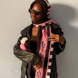 Puede incluir: Conjunto de gorro y bufanda de ganchillo en marr&oacute;n, blanco y rosa. La bufanda tiene un dise&ntilde;o estampado con detalles calados. El gorro es a rayas. La persona lleva gafas de sol y una chaqueta de cuero negra, sosteniendo lana.