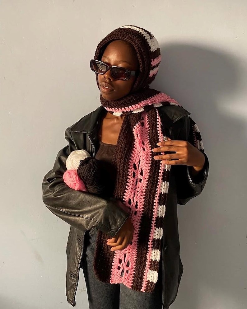 Puede incluir: Conjunto de gorro y bufanda de ganchillo en marr&oacute;n, blanco y rosa. La bufanda tiene un dise&ntilde;o estampado con detalles calados. El gorro es a rayas. La persona lleva gafas de sol y una chaqueta de cuero negra, sosteniendo lana.