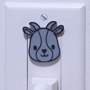 Peut inclure: Une prise électrique blanche avec un autocollant de visage de chèvre de dessin animé. La chèvre a une fourrure grise et un contour noir. Un cordon électrique blanc est branché dans la prise. La prise est fixée sur un mur blanc.