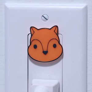 Puede incluir: Una cubierta blanca de enchufe con una pegatina de cara de zorro de dibujos animados. El zorro es naranja con contornos negros y un fondo blanco. Un cable eléctrico blanco está enchufado en el enchufe.