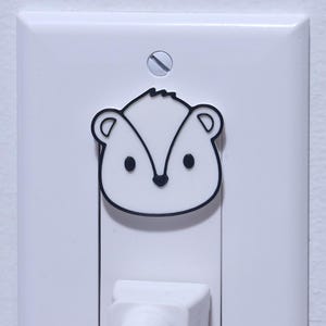 Puede incluir: Una cubierta blanca para tomacorrientes con una cubierta de enchufe en forma de mofeta. La mofeta es negra y blanca, con un diseño de dibujos animados. Un cable eléctrico blanco está enchufado en el tomacorriente.