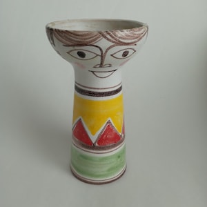 May include: A whimsical ceramic vase with a face design. The top features a smiling face with brown hair and pink cheeks. The body is decorated with yellow, red, and green geometric patterns. A unique decorative piece.