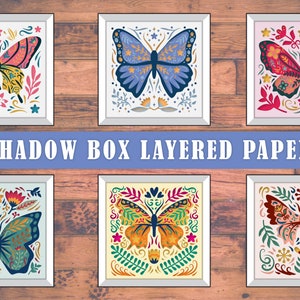May include: Six framed butterfly art prints with detailed designs in various colours, including pink, blue, and orange. The text "3D SHADOW BOX LAYERED PAPERCUT" is displayed across the image. The artwork is set against a wooden background.