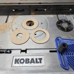 May include: Three wooden rings with different sized holes in the center. The rings are on a light gray surface with a black and silver tool and a blue plastic tool.