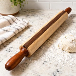 May include: A wooden rolling pin with a dark brown center stripe and matching handles. The rolling pin is on a countertop with flour and a ball of dough, suggesting a baking theme. A striped kitchen towel and a potted plant are also visible.