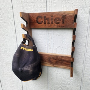 May include: A wooden hat rack with the word "Chief" engraved on the top. A navy blue baseball cap with the word "VETERAN" in yellow lettering is hanging on the rack. The rack is made of dark brown wood and is mounted on a white wall.