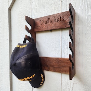 May include: A wooden hat rack with the words "Dad's hats" engraved on it. A black baseball cap with yellow lettering that says "Veteran" is hanging on the rack. The rack is mounted on a white wooden wall.