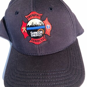 May include: A navy blue baseball cap with an embroidered patch featuring a red cross with a white circle in the centre. The circle has a cityscape and the text "Suncor Energy Services" in black. The cross has the text "Emergency" at the top and "Services" at the bottom.