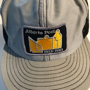 May include: A grey and black mesh trucker hat with a yellow and black patch that says "Alberta Pool 1973-1983".