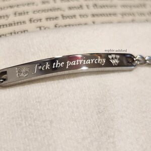 May include: A silver bracelet engraved with the words "f*ck the patriarchy". A butterfly and floral design are on either side of the text. The bracelet is displayed on a white surface.