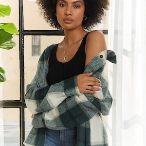May include: A woman wearing a black tank top and light blue jeans, with a green and white plaid flannel shirt draped over her shoulders. She is wearing a silver necklace with a cross pendant.