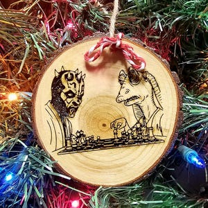 May include: Wooden Christmas ornament featuring a Star Wars theme. The ornament depicts Darth Maul and Jar Jar Binks playing chess. The artwork is burned into a round wooden slice, with a twine and red/white string for hanging.