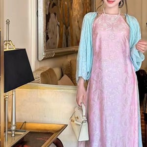 May include: A light pink sleeveless dress with a subtle tonal pattern, paired with a light blue long-sleeved bolero jacket and matching wide-leg trousers. The outfit is accessorized with a white handbag and a pearl necklace.