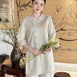 K&ouml;nnte beinhalten: Ein beiges Leinen-Outfit, bestehend aus einer Tunika mit Lotusblumenstickerei und passender Hose. Die Tunika hat einen Stehkragen und Kn&ouml;pfe. Die Person h&auml;lt wei&szlig;e Blumen. Der Hintergrund zeigt Bambus und eine sitzende Figur.