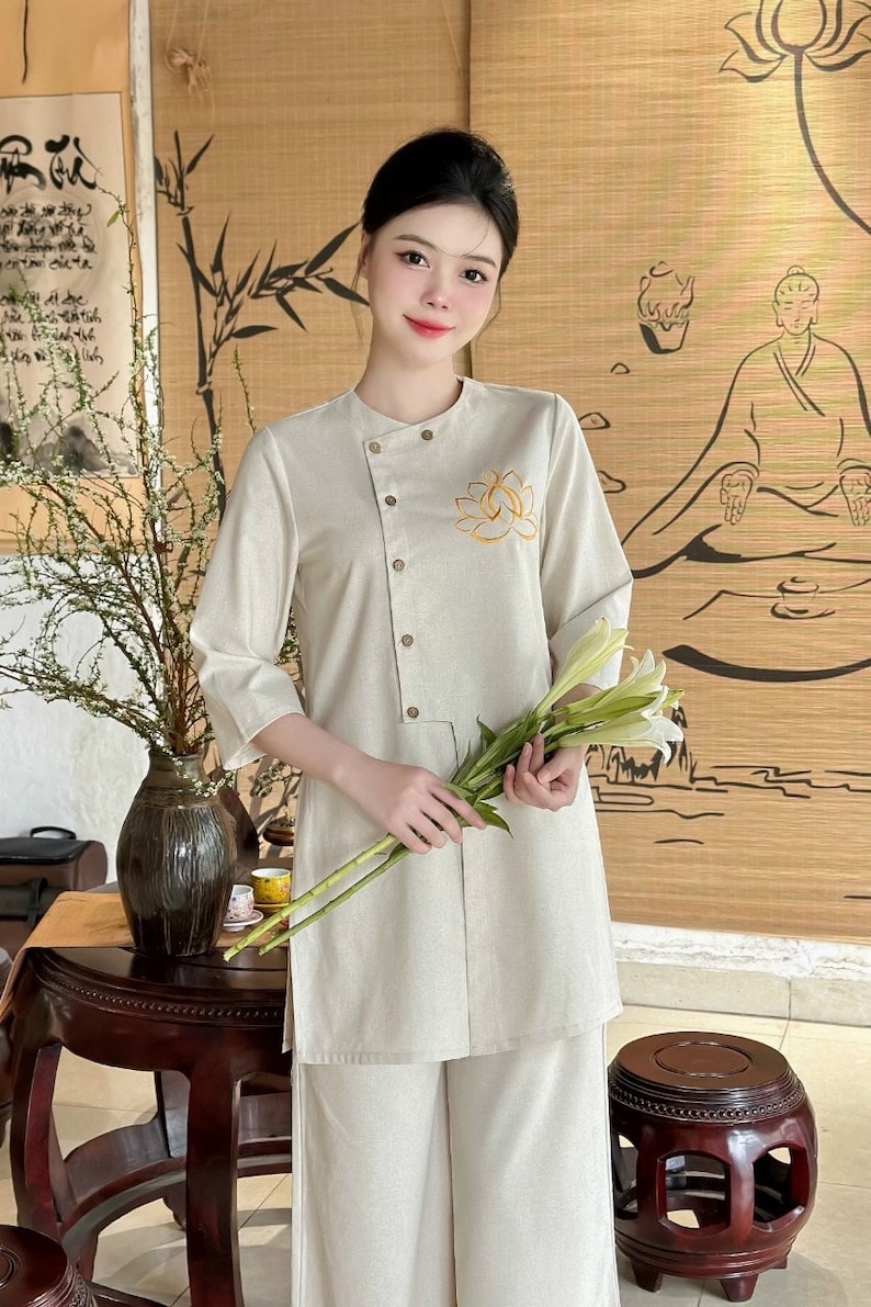 K&ouml;nnte beinhalten: Ein beiges Leinen-Outfit, bestehend aus einer Tunika mit Lotusblumenstickerei und passender Hose. Die Tunika hat einen Stehkragen und Kn&ouml;pfe. Die Person h&auml;lt wei&szlig;e Blumen. Der Hintergrund zeigt Bambus und eine sitzende Figur.