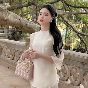 May include: A person wearing a cream-colored outfit, including a top with flared sleeves and matching pants. They are holding a pink quilted handbag with gold-colored hardware. The background features a stone wall and lush greenery.