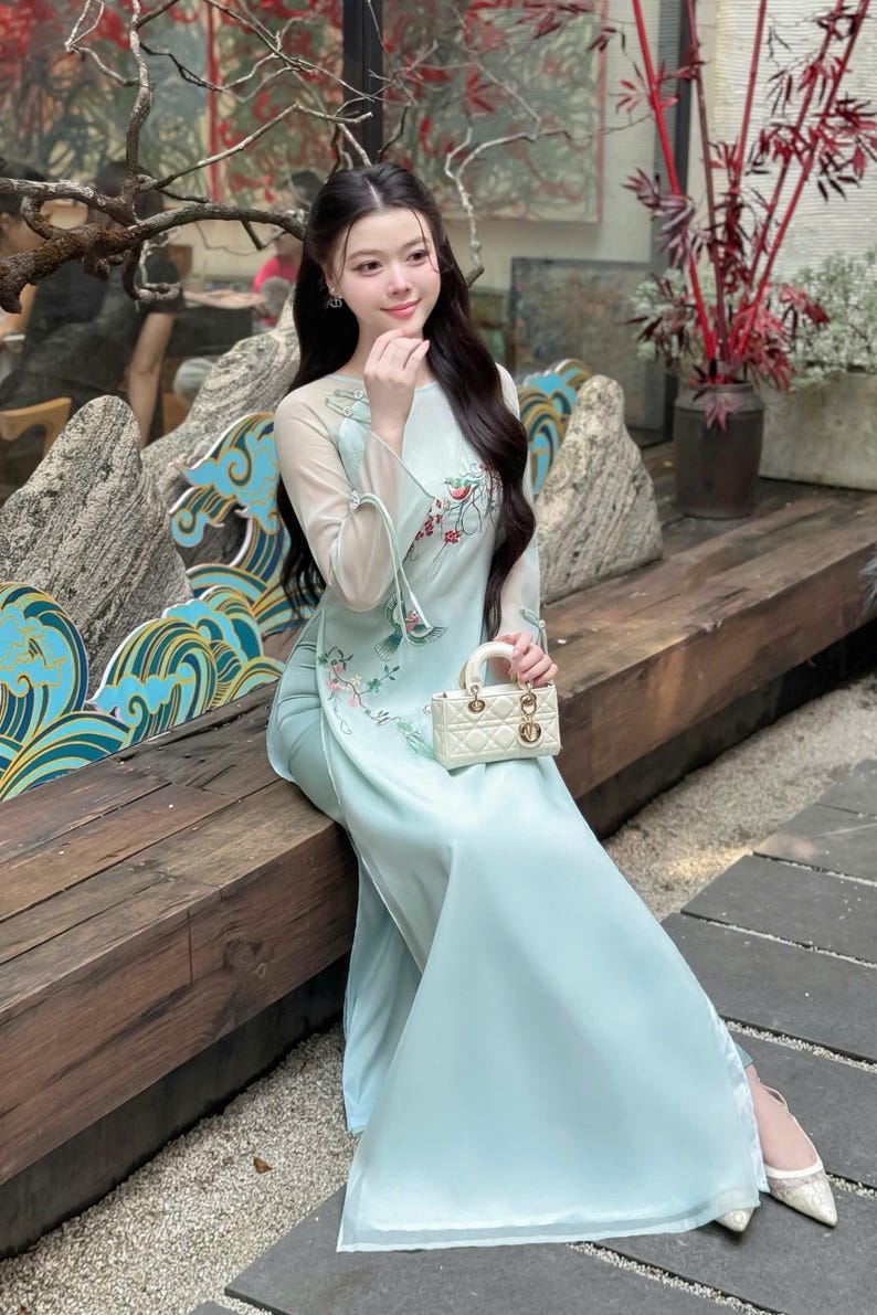 Puede incluir: Un vestido largo azul claro con bordados florales y mangas transparentes. El vestido se combina con un peque&ntilde;o bolso de mano blanco. La mujer est&aacute; sentada en un banco de madera al aire libre. El vestido tiene un cuello alto y una falda fluida.