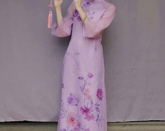 May include: A woman in a light purple floral dress and matching pants plays a wooden flute. The dress features a floral pattern in shades of purple and pink. The outfit is complemented by a matching light purple pants. The background is a solid purple.