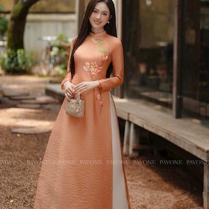 May include: A long, peach-colored dress with floral embroidery and long sleeves. The dress has a high collar and a split front, revealing a white underlayer. A small, patterned handbag is held in the hand.