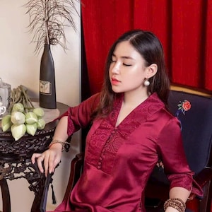 May include: A burgundy silk tunic and matching pants. The tunic features a V-neck with lace detailing and button accents. The sleeves are three-quarter length. The outfit is worn with beaded bracelets. A black fan is held in the hand.