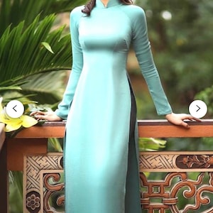 May include: A woman wearing a light blue, long-sleeved dress with a high collar and a side slit. The dress is made of a silky fabric and has a fitted silhouette. The woman is standing in front of a wooden railing with intricate carvings.