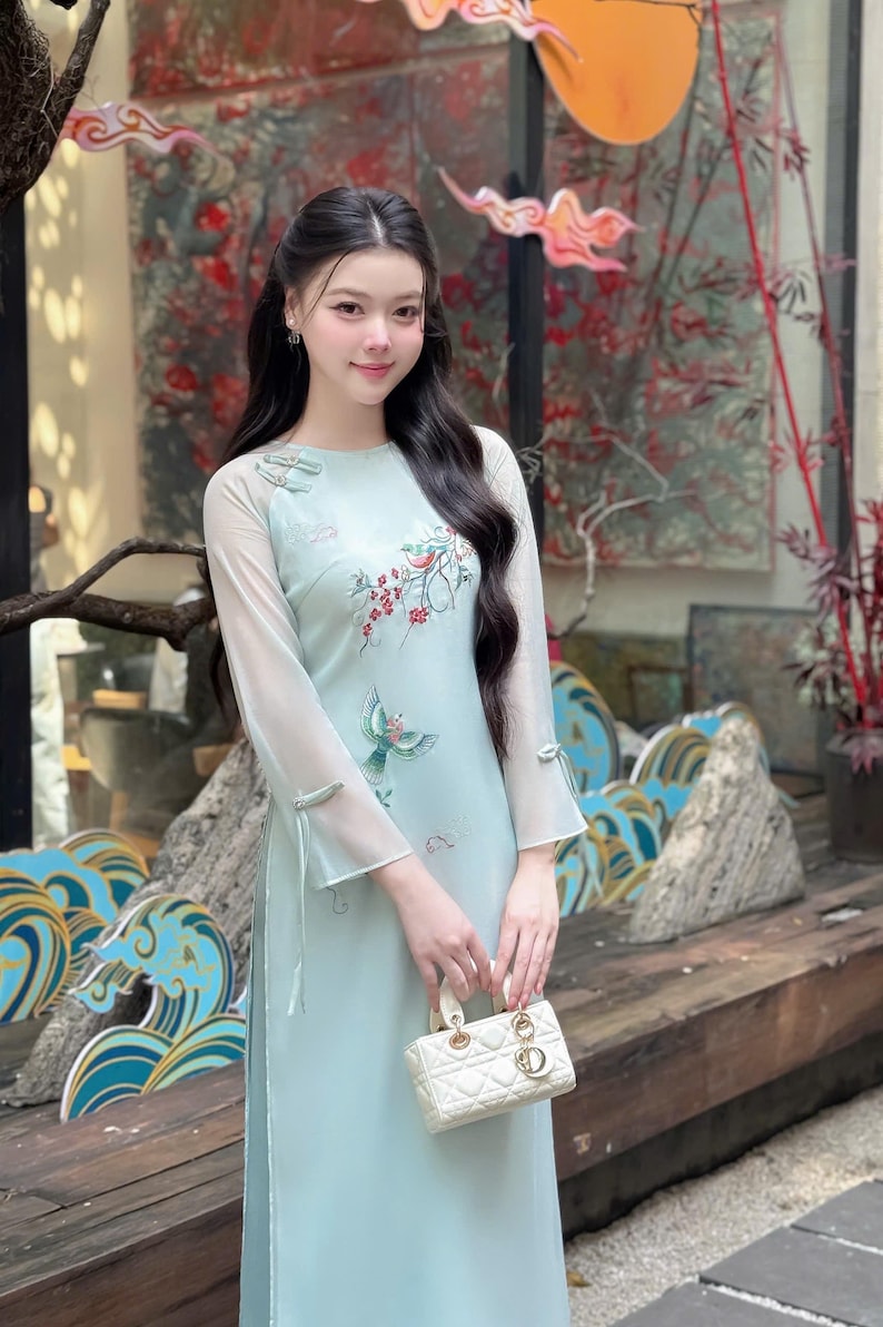 Puede incluir: Un vestido azul claro con mangas largas y transparentes y bordados florales. El vestido presenta un cuello redondo y un peque&ntilde;o bolso de mano blanco. El estilo es elegante y tradicional, adecuado para ocasiones especiales.