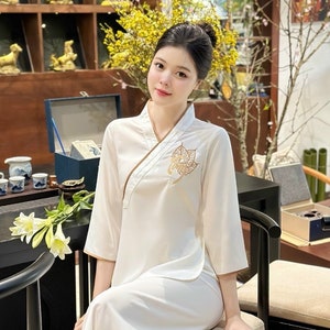 May include: A woman wearing a white, long-sleeved outfit with gold trim and a leaf design on the front. The outfit includes matching pants. She is seated in a black chair. The background includes yellow flowers and decorative items.
